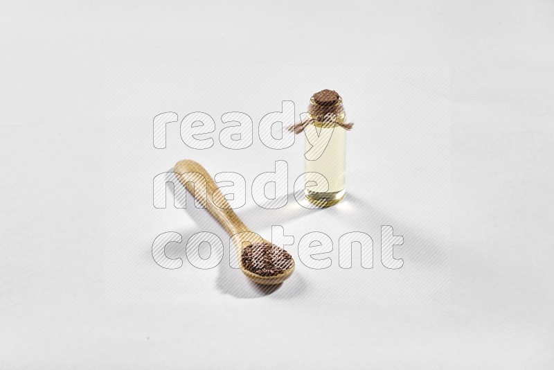 A wooden spoon full of flax seeds and a glass bottle full of flaxseeds oil on a white flooring