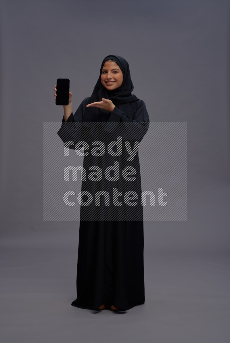 Saudi woman wearing Abaya standing showing phone to camera on gray background