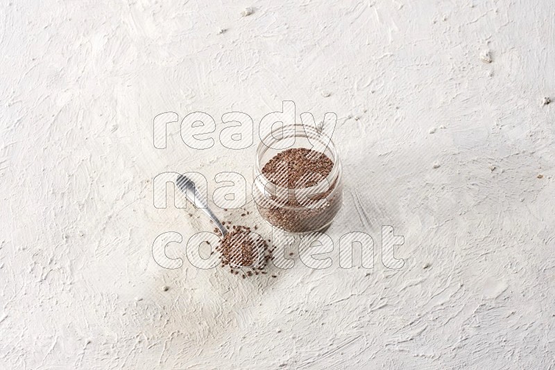A glass jar full of flax and a metal spoon full of the seeds on a textured white flooring in different angles