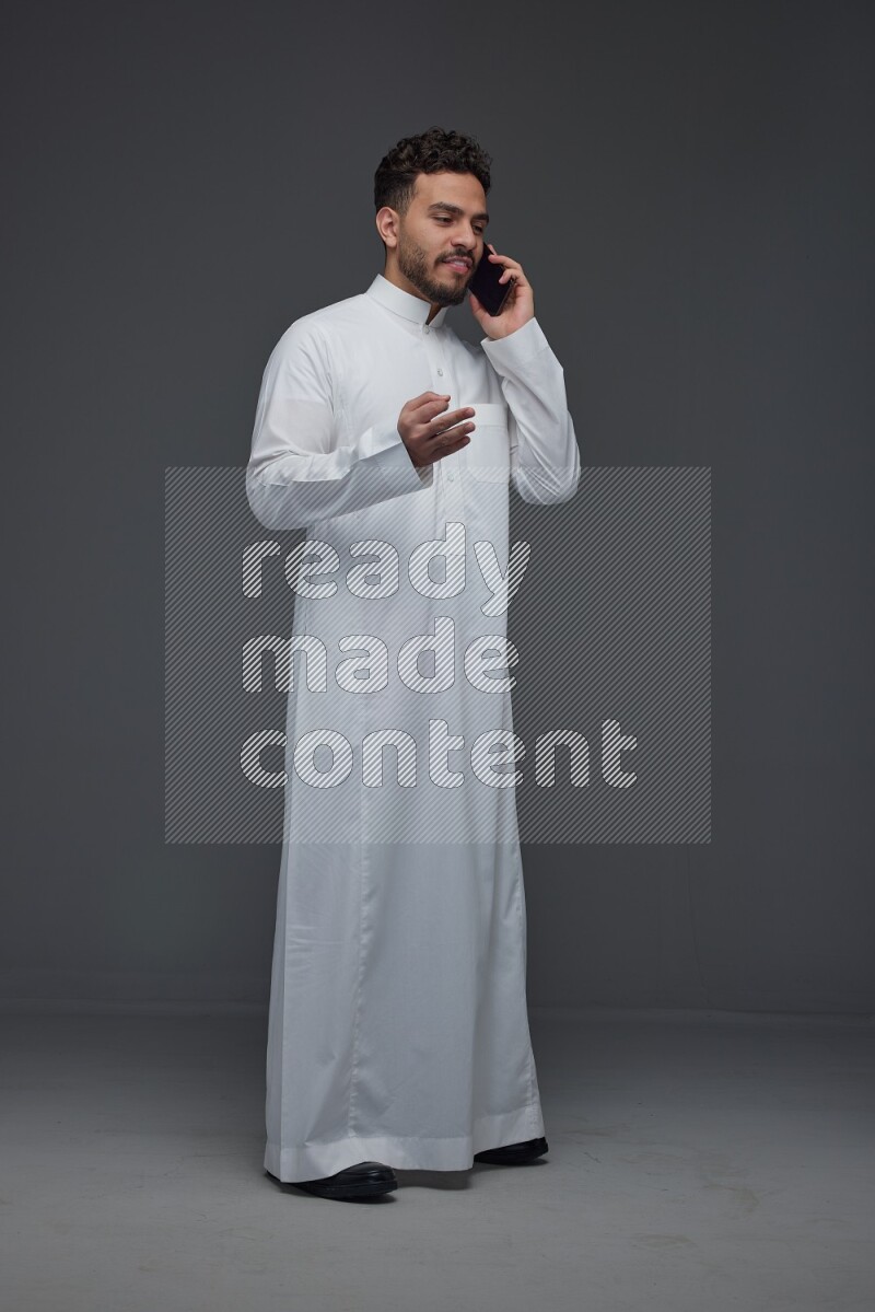 A Saudi man wearing Thobe and talking in the phone while standing and making different poses eye level on a gray background
