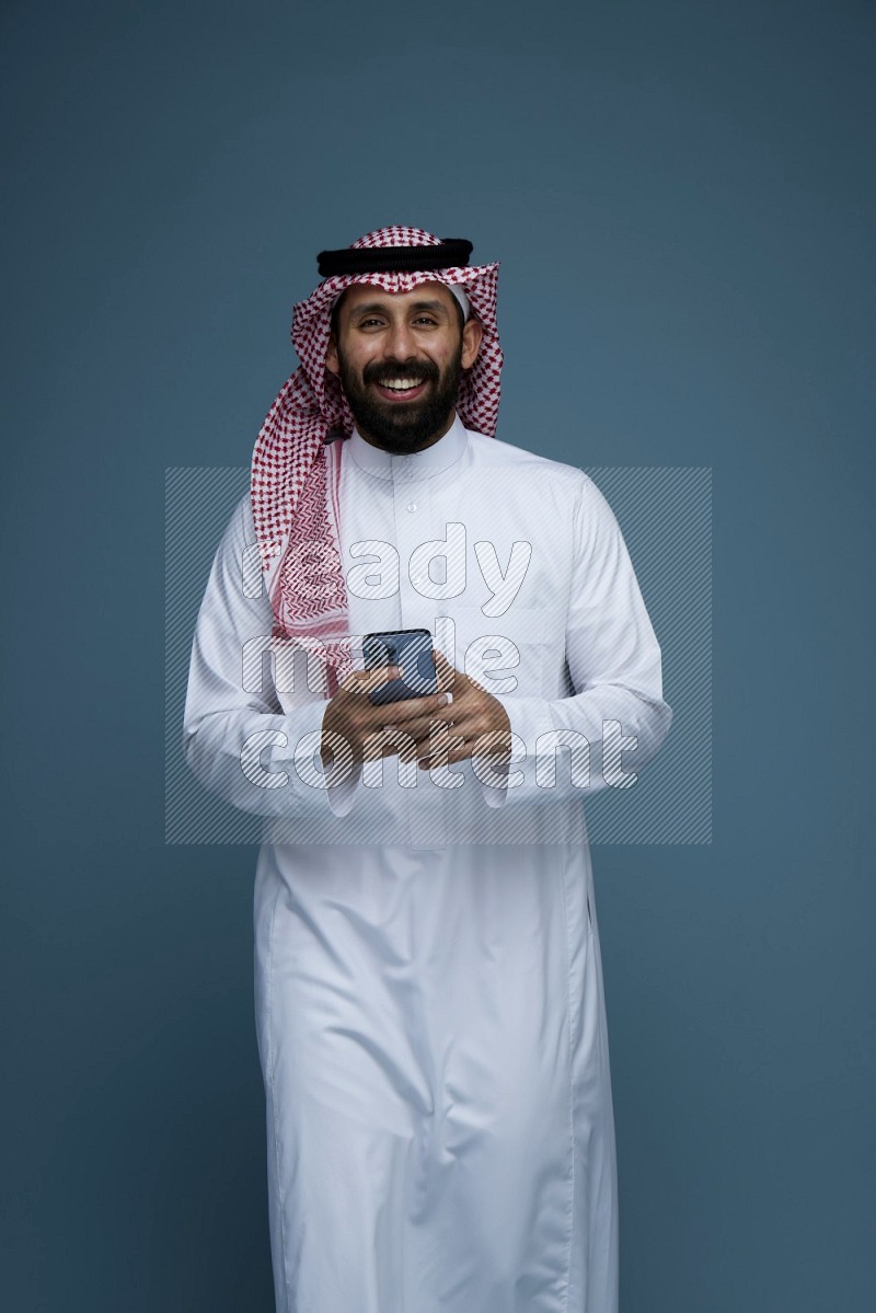 A Male posing with a phone in a blue background wearing Saudi Thob and Shomag