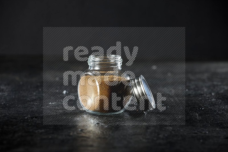 A glass spice jar full of cumin powder on a textured black flooring