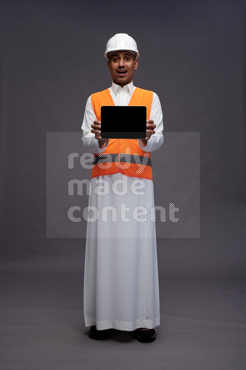Saudi man wearing thob with engineer vest standing showing tablet to camera on gray background