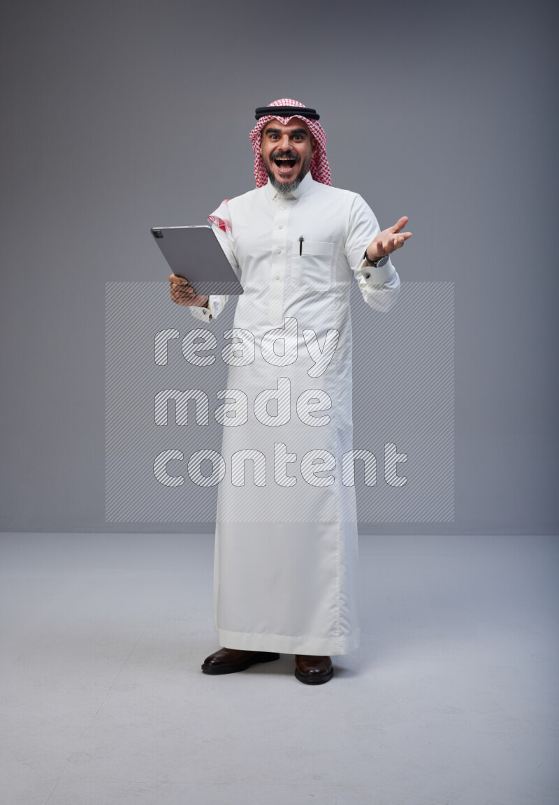 Saudi man Wearing Thob and red Shomag standing working on tablet on Gray background