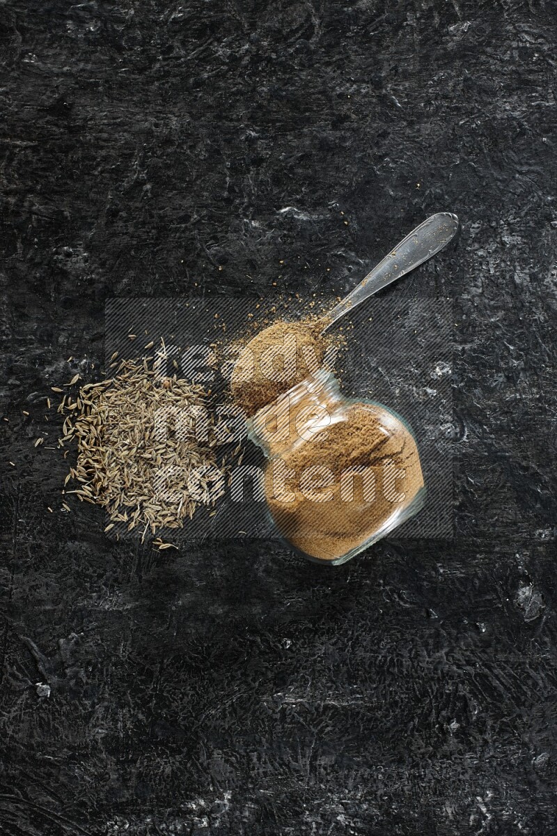 A flipped glass spice jar and a metal spoon full of cumin powder and powder spilled out with cumin seeds on a textured black flooring