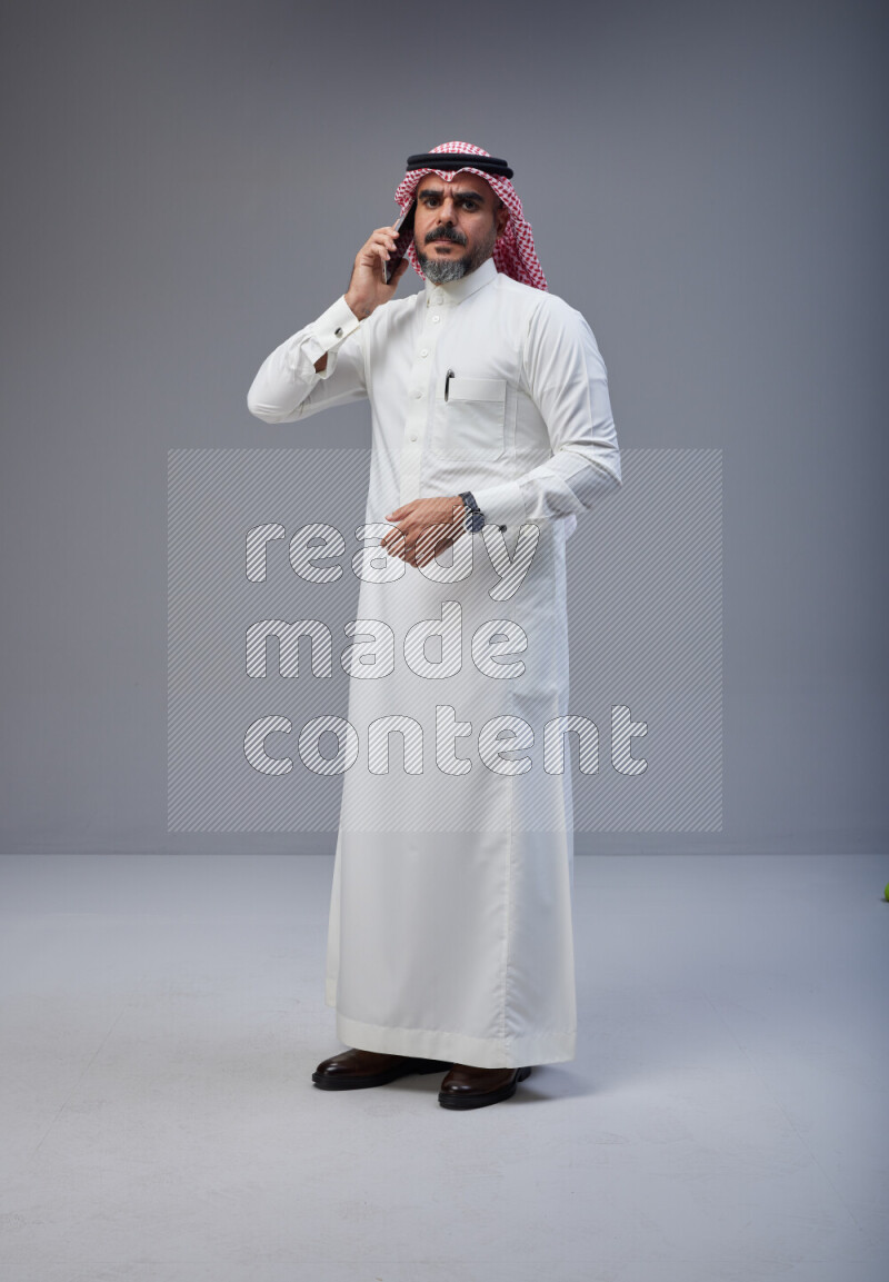 Saudi man Wearing Thob and red Shomag standing talking on phone on Gray background