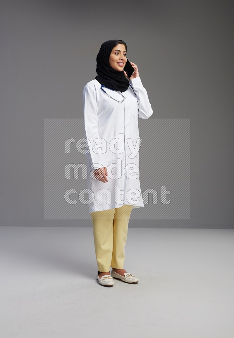 Saudi woman wearing lab coat with stethoscope standing talking on phone on Gray background