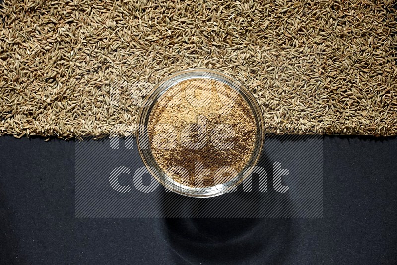 A glass bowl full of cumin powder and cumin seeds beneath it on black flooring
