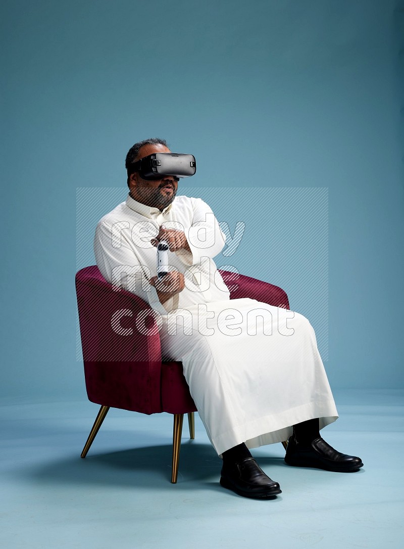 Saudi Man without shimag sitting on chair  gaming with VR and joystick on blue background