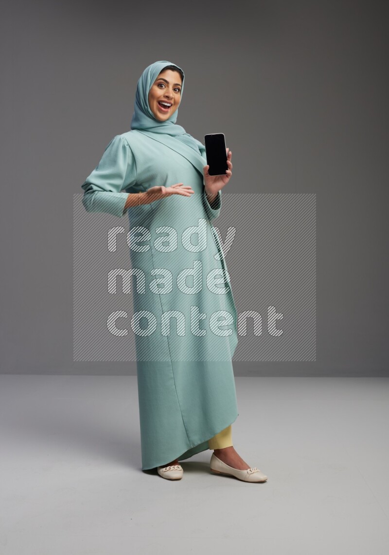 Saudi Woman wearing Abaya standing showing phone to camera on Gray background