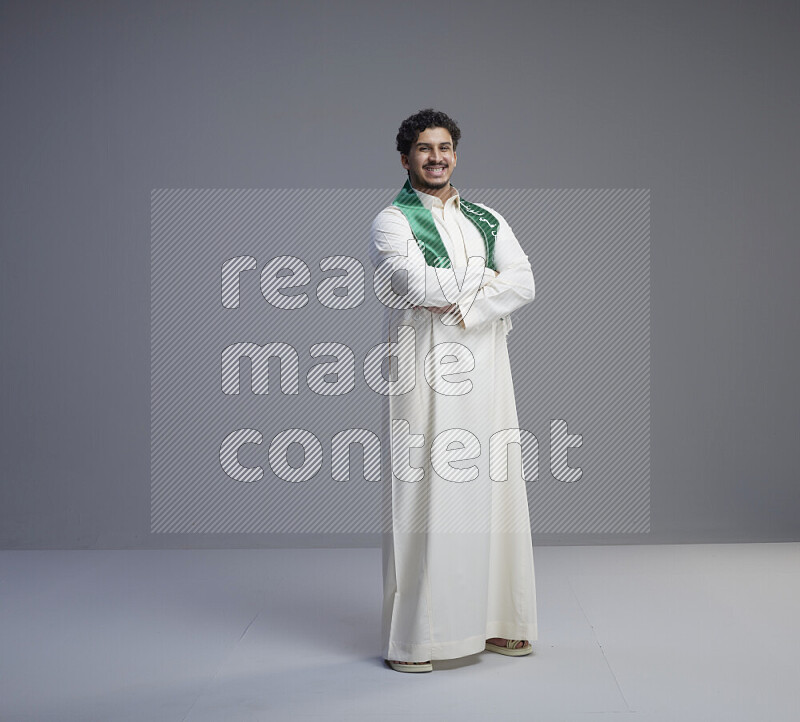 A Saudi man standing wearing thob and Saudi flag scarf on gray background