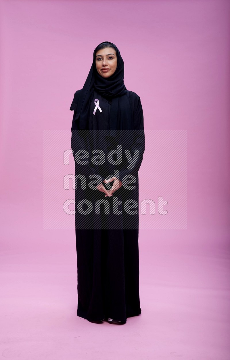 Saudi woman wearing pink ribbon on Abaya standing interacting with the camera on pink background