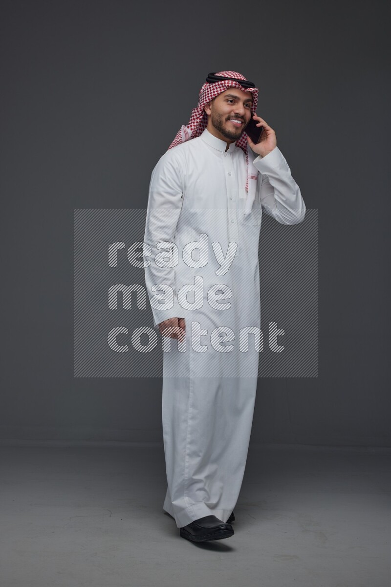 A Saudi man wearing Thobe and Shmagh talking in the phone eye level on a gray background