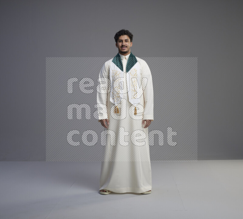 A Saudi man standing wearing thob and Saudi flag scarf on gray background