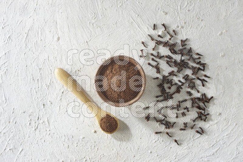 A wooden bowl and wooden spoon full of cloves powder with cloves spread on textured white flooring