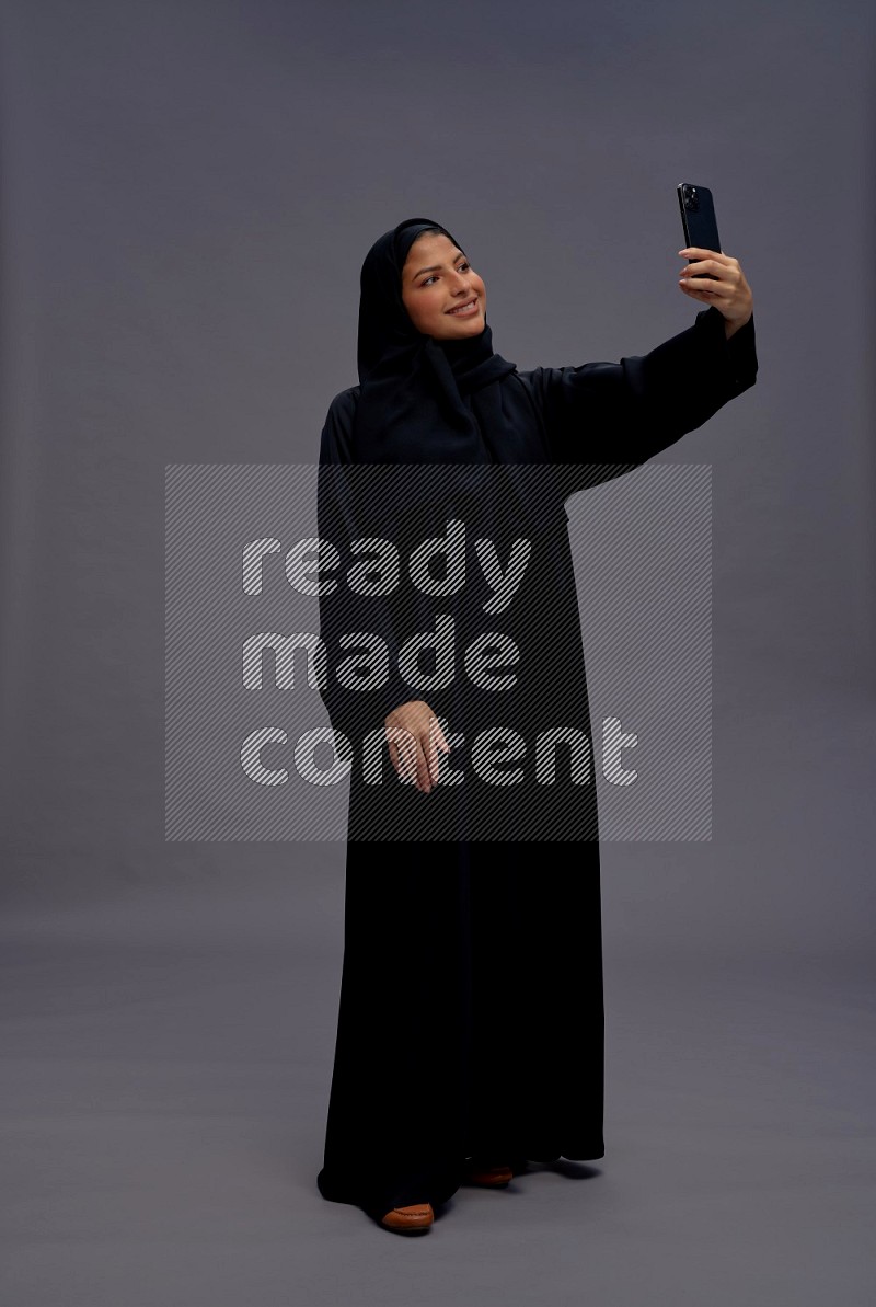 Saudi woman wearing Abaya standing taking selfie on gray background