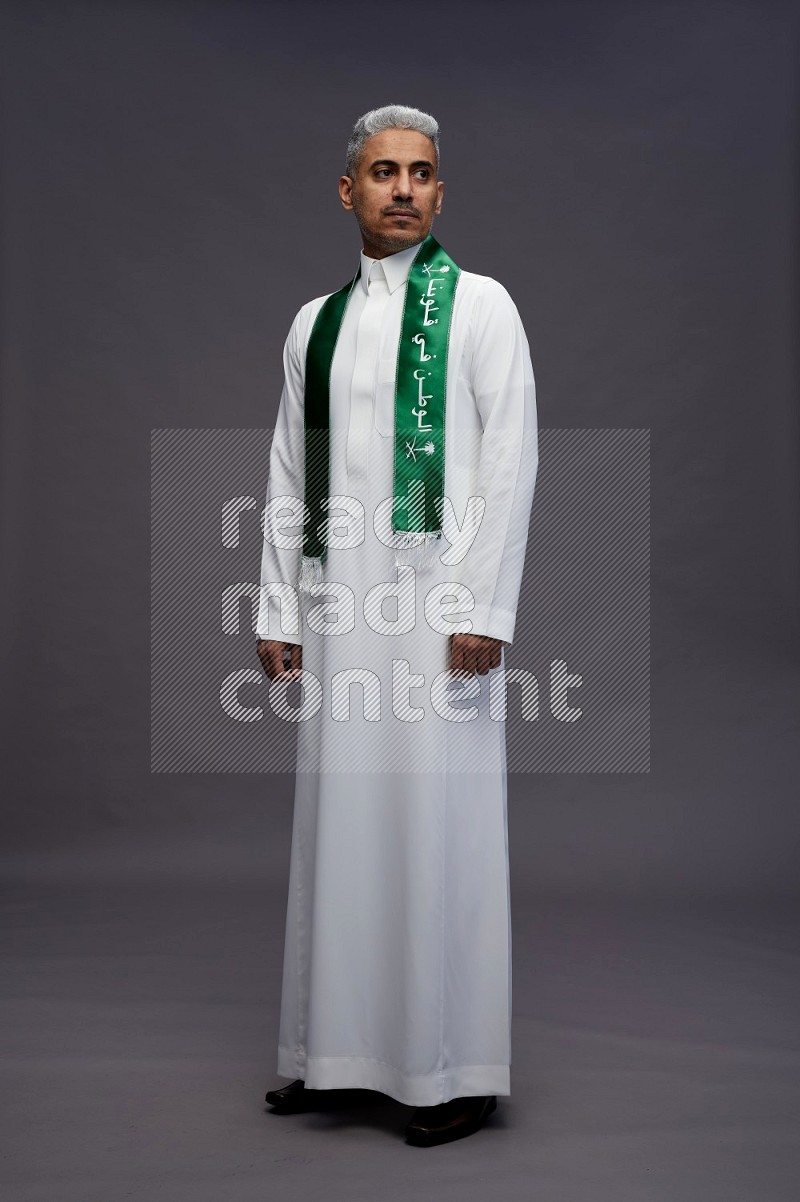 Saudi man wearing thob standing holding Saudi flag on gray background