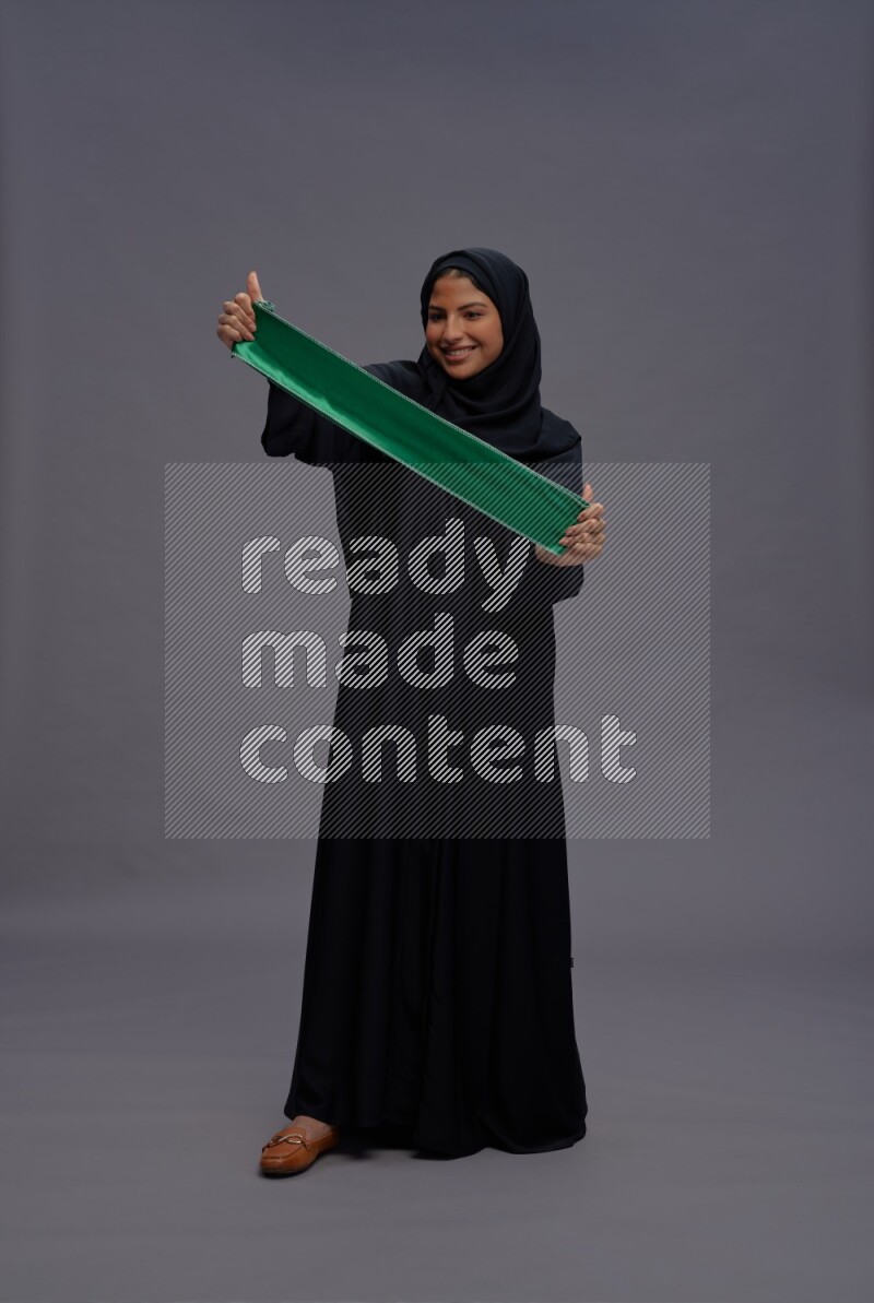 Saudi woman wearing Abaya standing holding Saudi flag on gray background