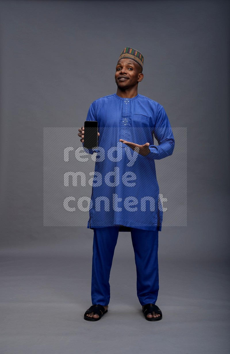 Man wearing Nigerian outfit standing showing phone to camera on gray background