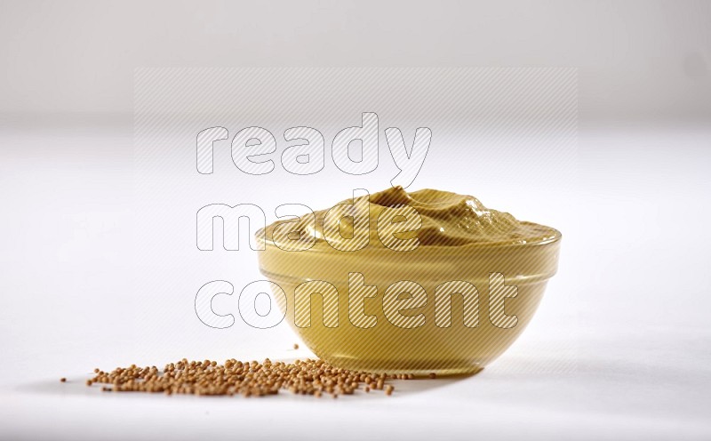 A glass bowl full of mustard paste with mustard seeds underneath on white flooring