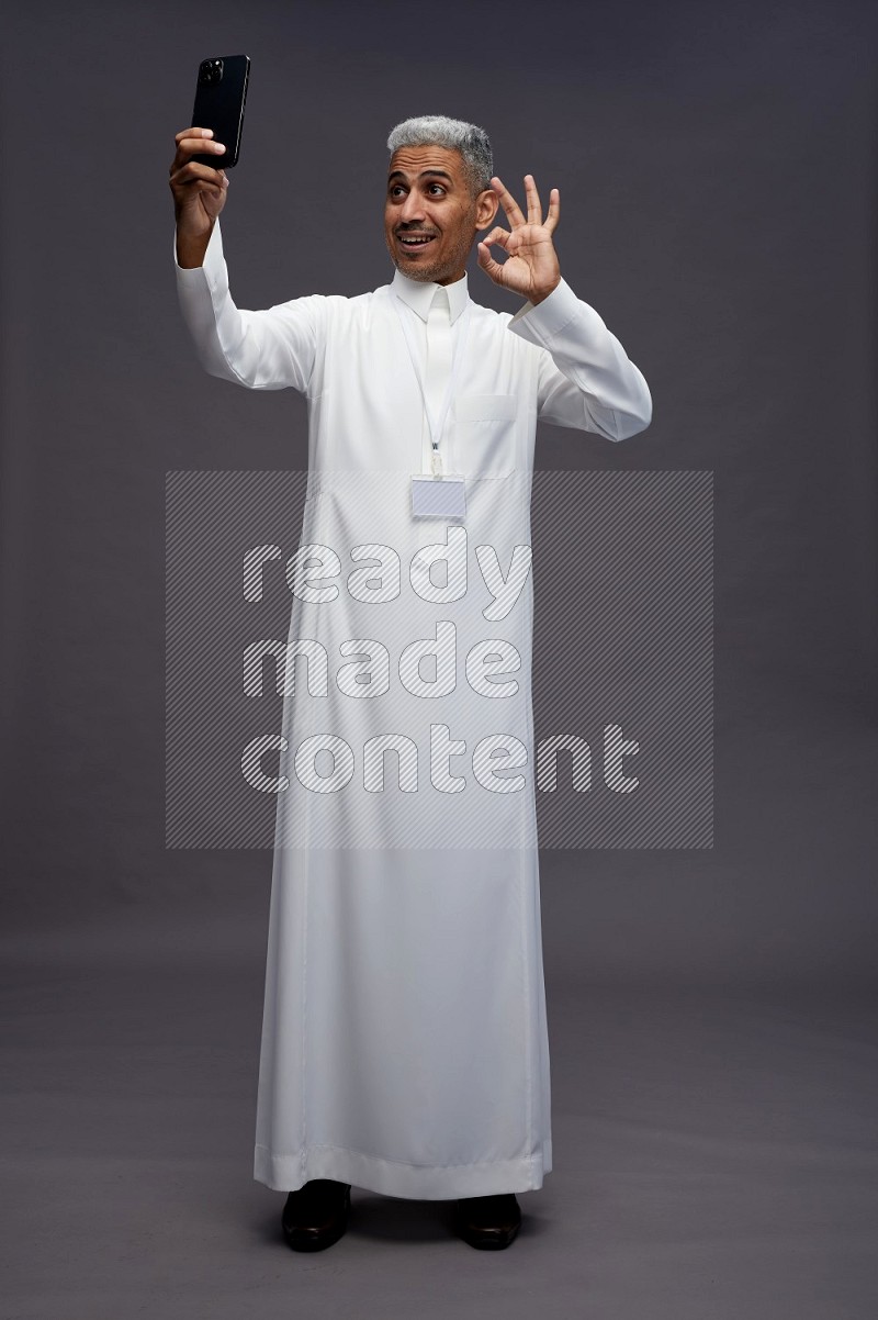 Saudi man wearing thob with neck strap employee badge standing taking selife on gray background