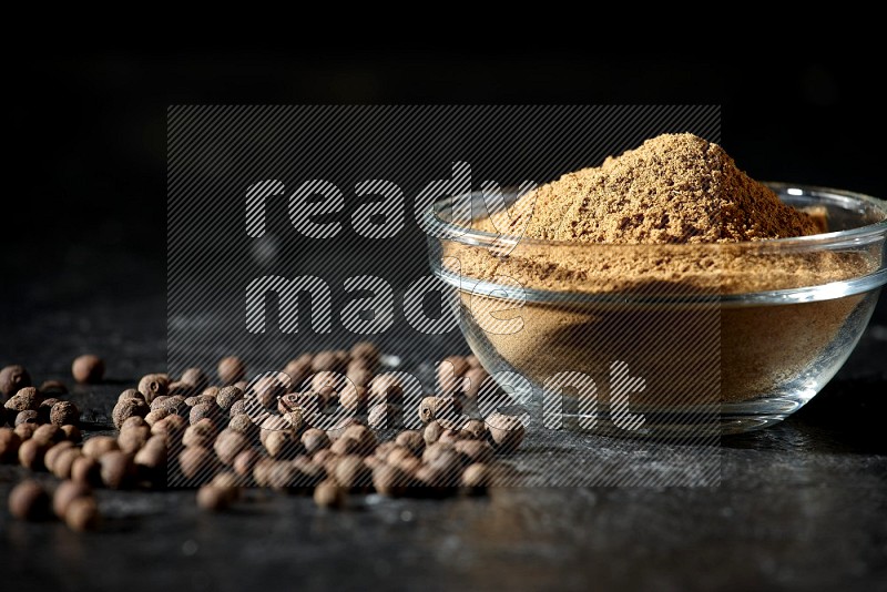 A glass bowl full of allspice powder and whole balls spreaded on a black flooring