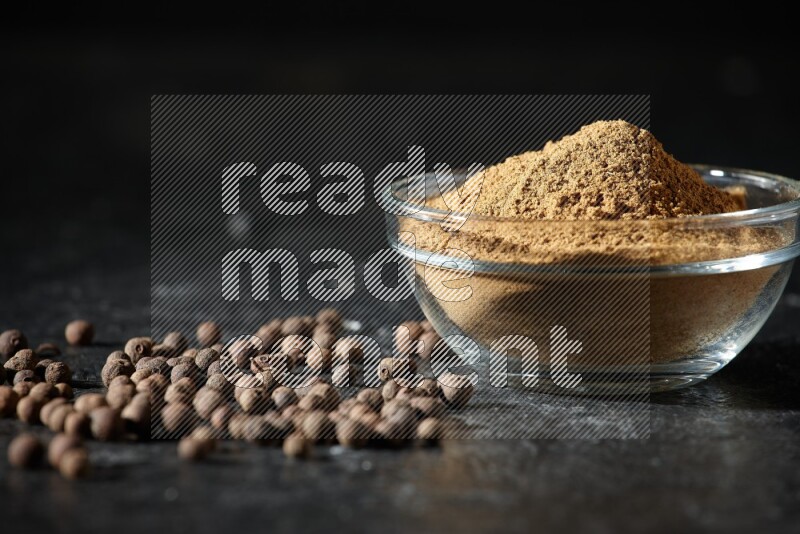 A glass bowl full of allspice powder and whole balls spreaded on a black flooring