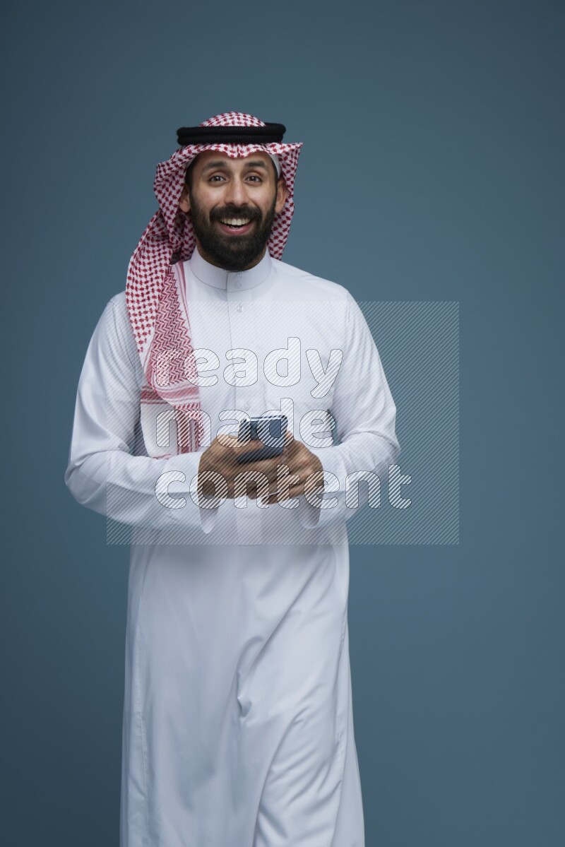 A Male posing with a phone in a blue background wearing Saudi Thob and Shomag