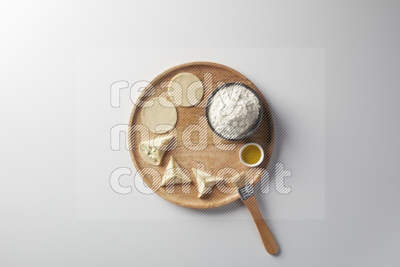 two closed sambosas and one open sambosa filled with cheese while flour, and oil with oil brush aside in a wooden dish on a white background