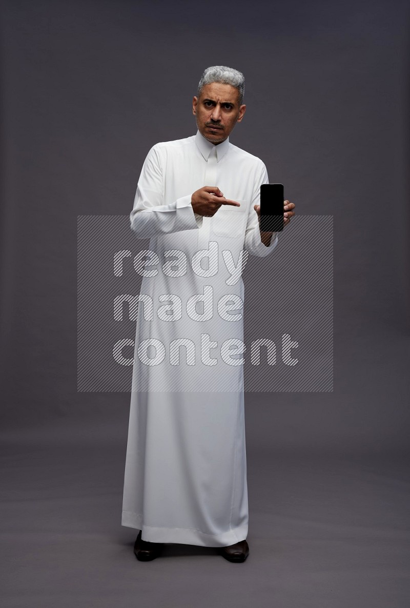 Saudi man wearing thob standing showing phone to camera on gray background