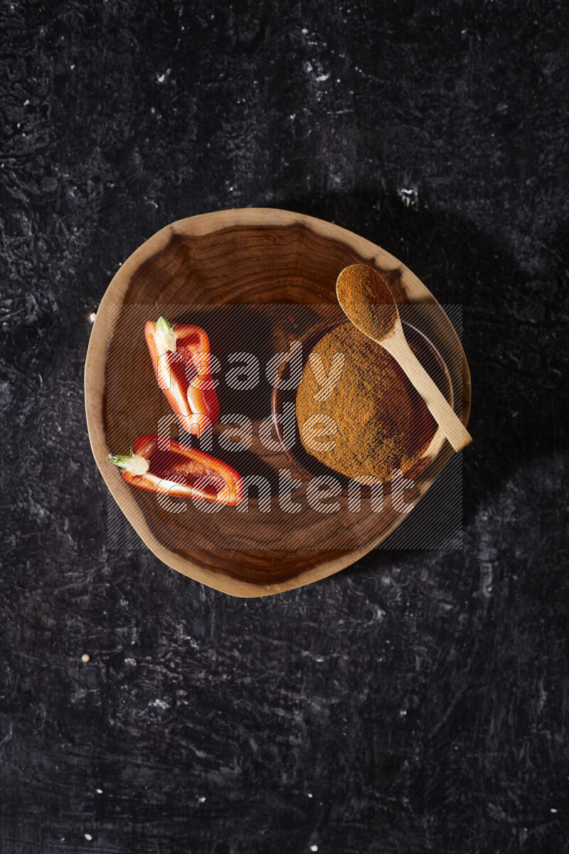 A wooden bowl full of ground paprika powder and sliced red bell pepper beside it, all on a wooden tray on black background