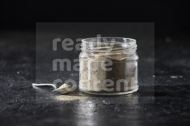 A glass jar and a metal spoon full of white pepper powder on textured black flooring