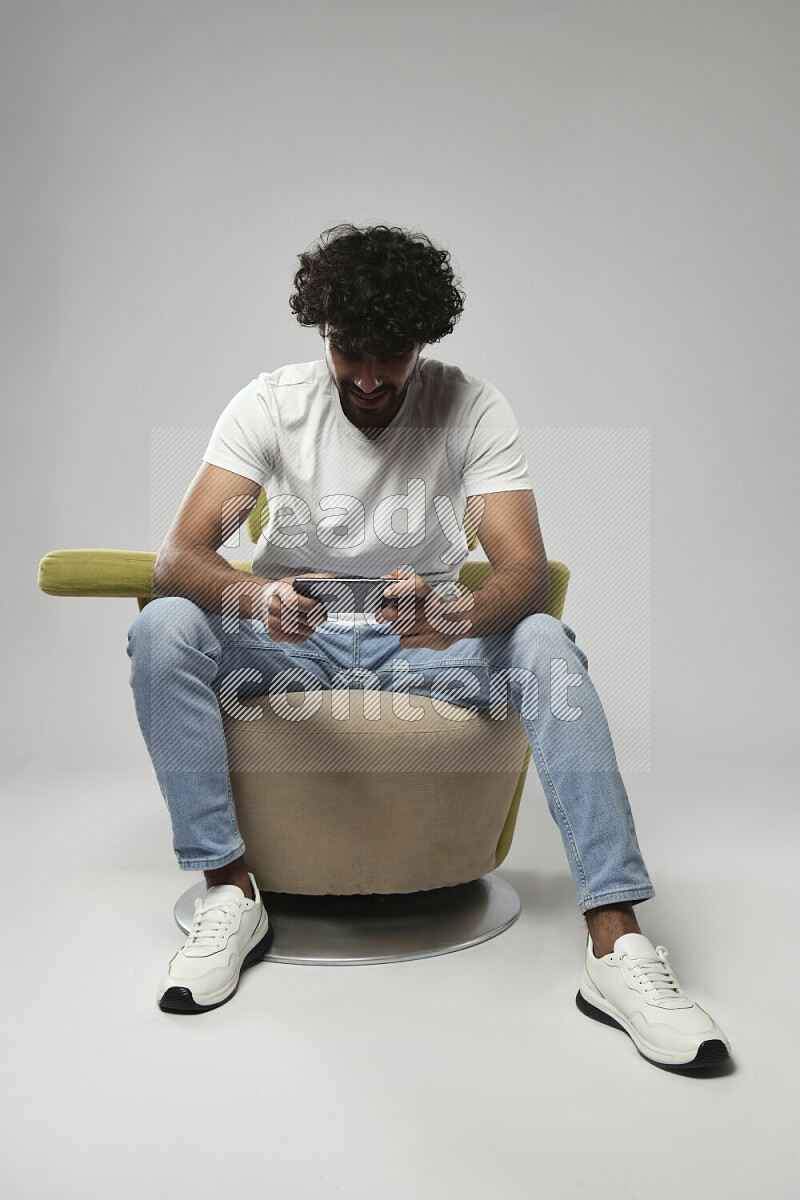رجل يرتدي ملابس كاجوال يجلس علي كرسي و يلعب علي الهاتف علي خلفية بيضاء