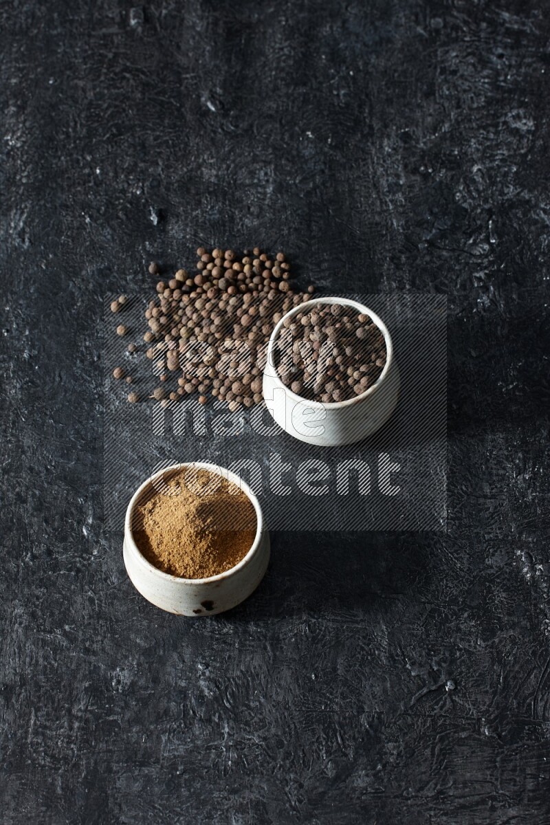 2 beige bowls, one full of allspice powder and the other full of whole balls and both spread on a textured black flooring