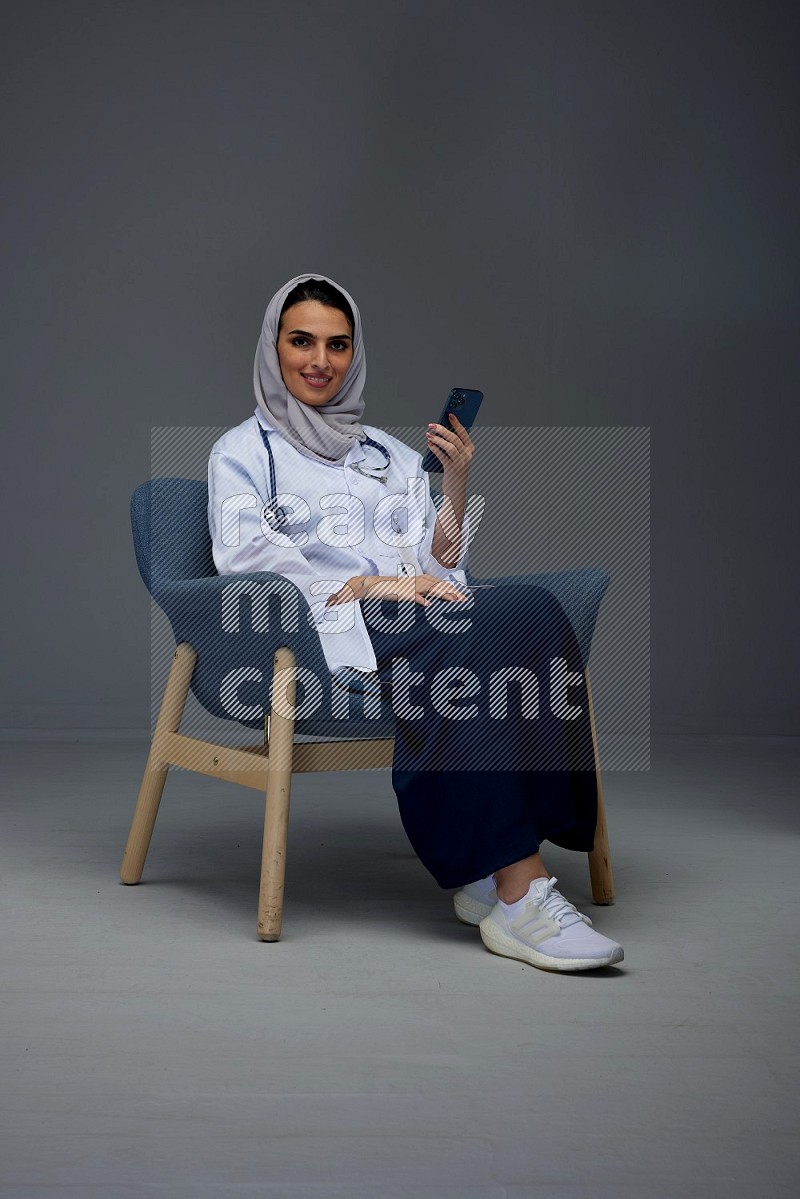 A doctor wearing a light gray head scarf sitting on a dark grey chair and using a laptop while talking in the phone eye level on a grey background