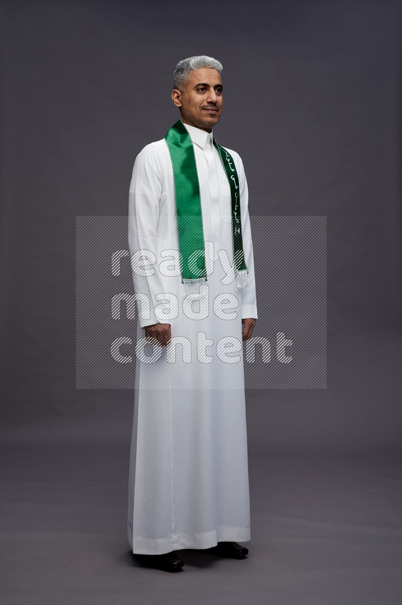 Saudi man wearing thob standing holding Saudi flag on gray background