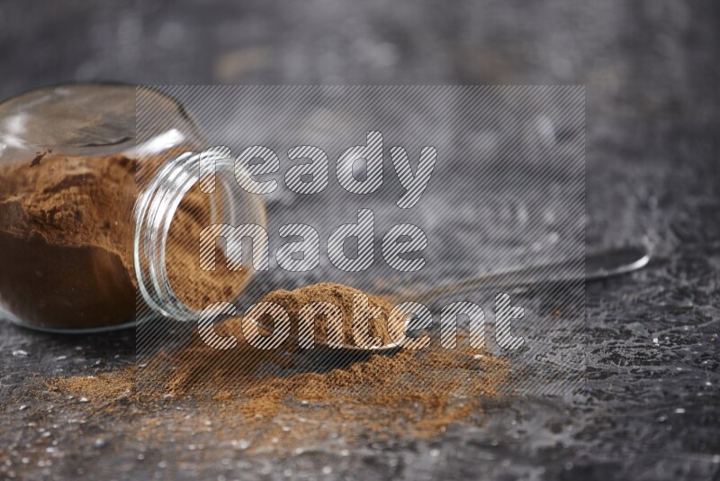 Herbal glass jar full of cinnamon powder flipped and a metal spoon on textured black background