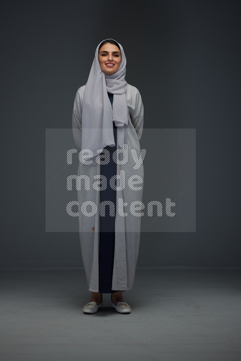 A Saudi woman wearing a light gray Abaya and head scarf standing and making multi poses eye level on a grey background