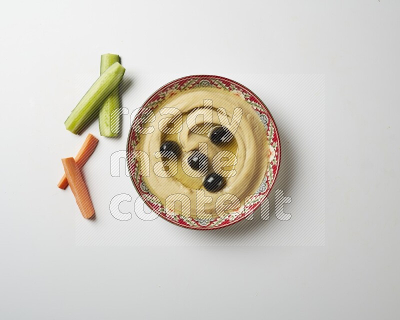 Hummus in a red plate with patterns garnished with black olives on a white background