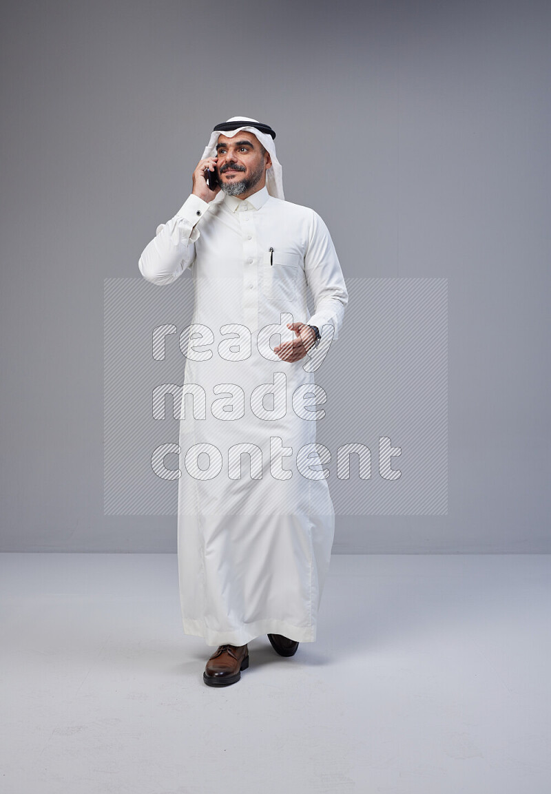 Saudi man Wearing Thob and white Shomag standing talking on phone on Gray background