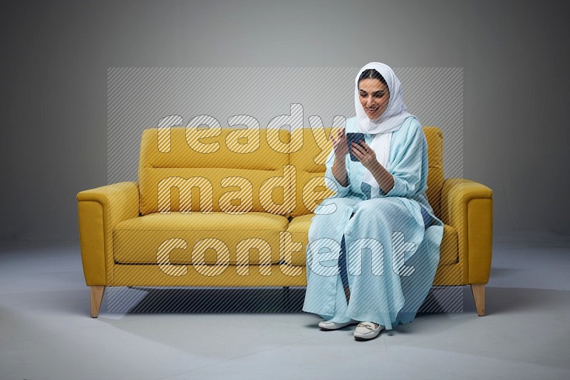 A Saudi woman wearing a light blue Abaya and white head scarf sitting on a yellow sofa and texting on her phone on a grey background