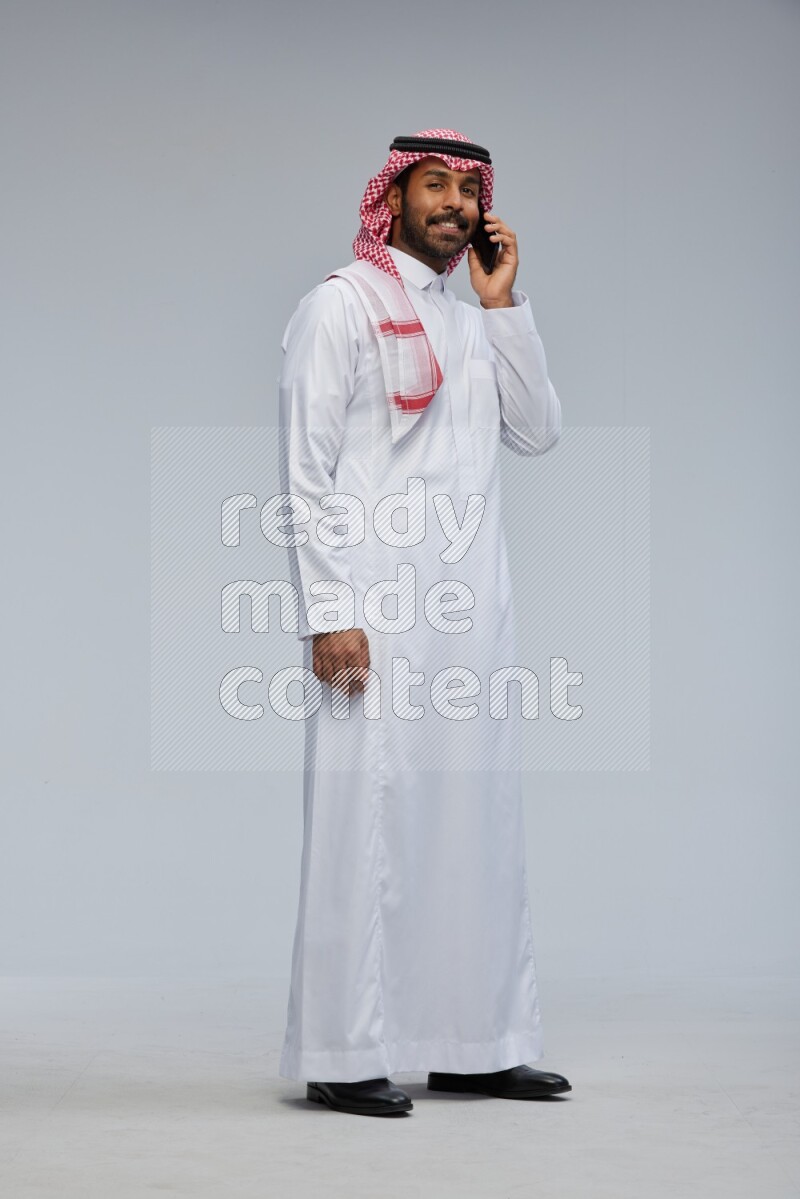 Saudi man Wearing Thob and shomag standing talking on phone on Gray background