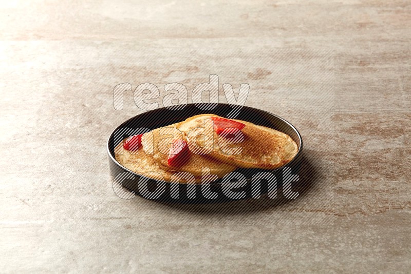 Three stacked strawberry pancakes in a black plate on beige background
