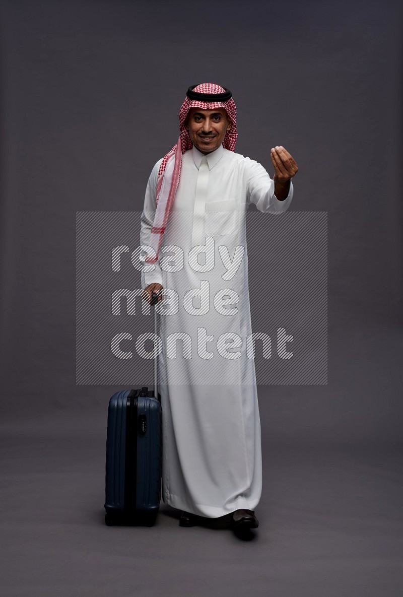 Saudi man wearing thob and shomag standing holding bag on gray background