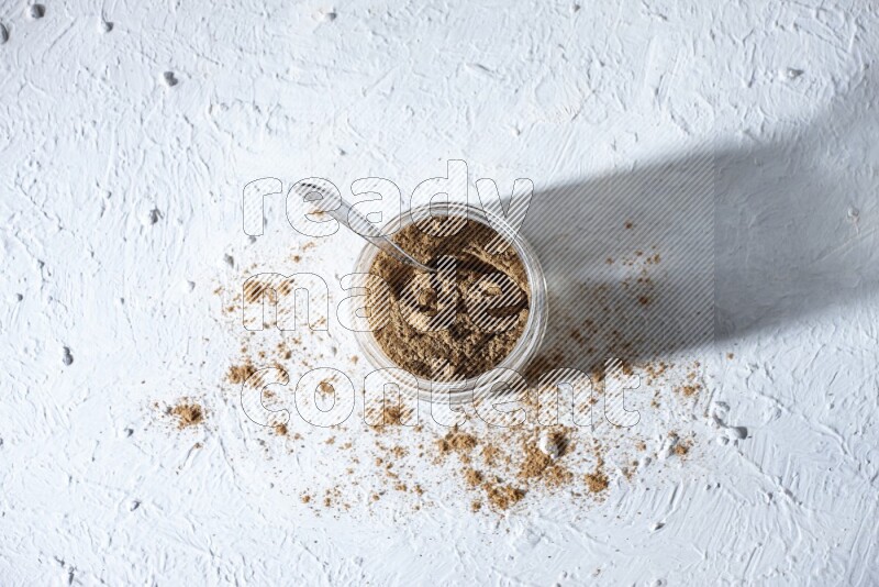 A glass jar and a metal spoon full of allspice powder on a textured white flooring