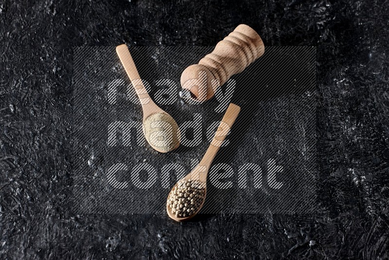 ملعقتان خشبيتان، واحدة ممتلئة ببودرة الفلفل الأبيض والأخرى بحبوب الفلفل ومطحنة خشبية على أرضية سوداء