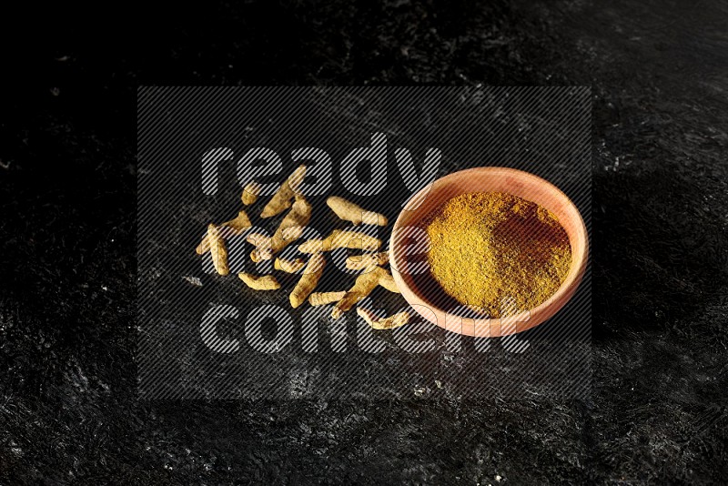 A wooden bowl full of turmeric powder with dried turmeric fingers on textured black flooring