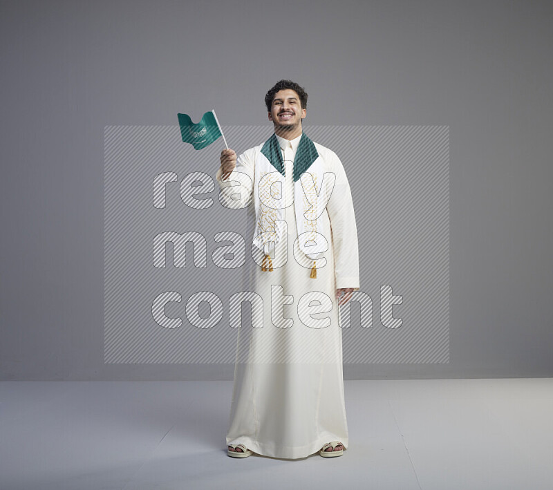 A Saudi man standing wearing thob and saudi flag scarf and holding small saudi flag on gray background