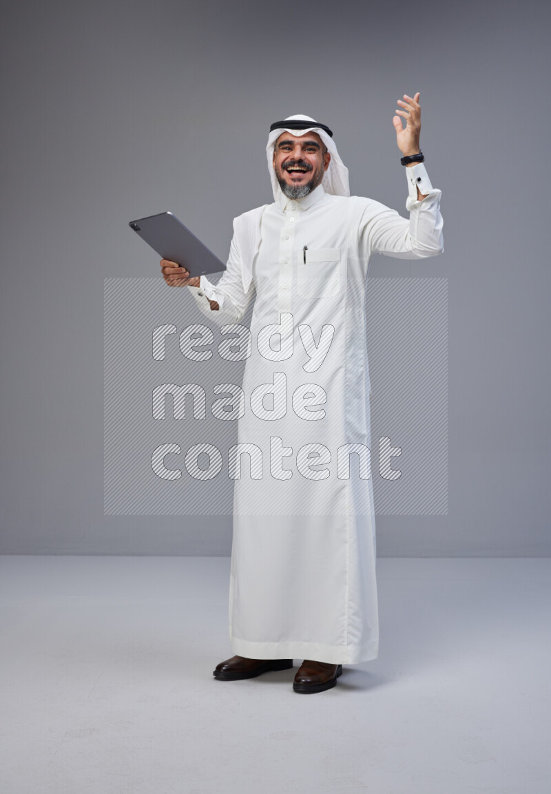 Saudi man Wearing Thob and white Shomag standing working on tablet on Gray background