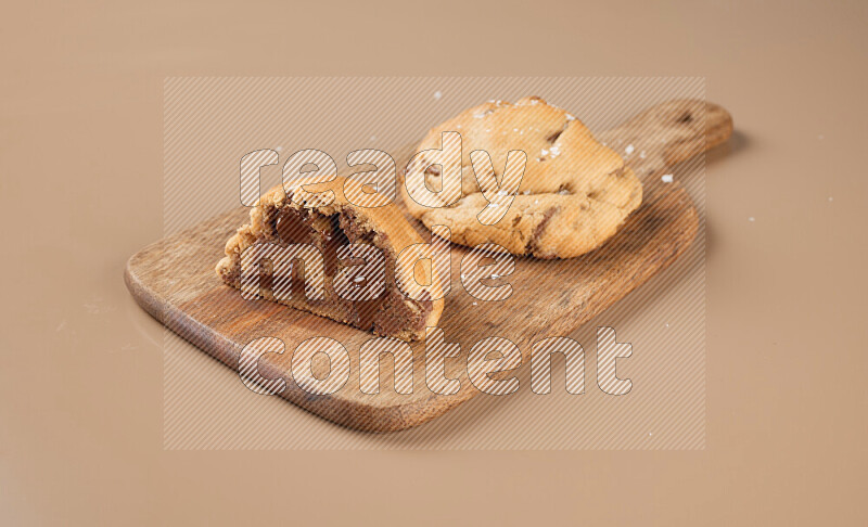 a chocolate chip cookie with another one cut in half on a wooden cutting board on a brown background